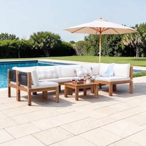 May include: An outdoor sectional sofa set with a wooden frame, white cushions, and black woven back accents. The set includes a low square coffee table and a large beige patio umbrella, all arranged on a stone patio beside a swimming pool.