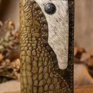 Pode incluir: Uma carteira retangular com um exterior de couro verde-oliva texturizado com um padrão de crocodilo. Um painel central de pele com pelo branco e castanho está fixado com rebites de latão. Um botão decorativo de metal escuro está centrado.