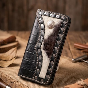 May include: A rectangular leather wallet featuring a black crocodile-embossed leather side and a central panel of white and dark brown cowhide hair-on leather. The wallet is accented with silver-toned metal studs and a circular silver-toned concho.