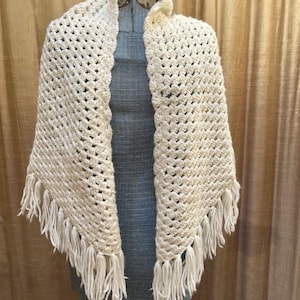 May include: A cream-colored triangular shawl featuring a textured crochet stitch pattern throughout the body. The edges of the shawl are finished with long, dangling fringe tassels that hang down from the bottom points of the garment.