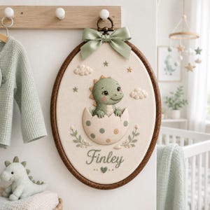 May include: An oval embroidery hoop wall hanging featuring a cream-colored fabric background with a stitched green dinosaur hatching from a polka-dot egg. The name Finley is embroidered in cursive below the dinosaur, accented by clouds, stars, and a sage green ribbon bow at the top.