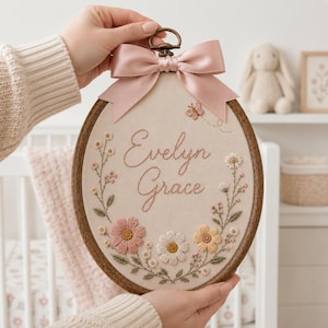 May include: An oval wooden embroidery hoop containing beige fabric with the embroidered name Evelyn Grace in cursive script. The design features a floral wreath with pink, white, and yellow flowers, green leaves, and a small butterfly, topped with a pink satin bow.