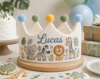 May include: A cream-colored felt crown featuring embroidered jungle animals including an elephant, zebra, lion, and giraffe. The name Lucas is stitched in blue script in the center. The crown is topped with five fuzzy pom-poms in blue, green, and yellow.