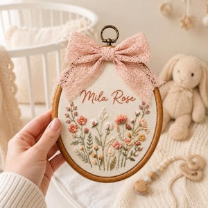 May include: An oval wooden embroidery hoop containing white fabric with the embroidered name Mila Rose in cursive script above a colourful floral arrangement of small pink, orange, and white flowers with green stems, topped with a pink lace bow.