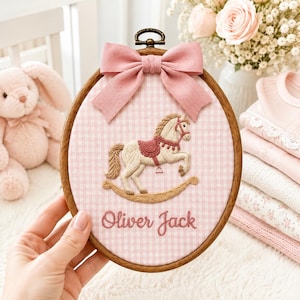 May include: An oval wooden embroidery hoop containing light pink and white gingham fabric. The fabric features an embroidered tan rocking horse with a dark pink saddle and the name Oliver Jack in cursive script, topped with a pink fabric bow.