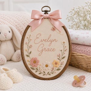 May include: An oval wooden embroidery hoop containing cream-colored fabric with the embroidered name Evelyn Grace in cursive script. The design features a floral wreath with pink, white, and yellow flowers, a small butterfly, and a pink satin bow.