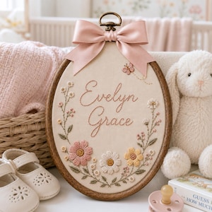 May include: An oval wooden embroidery hoop featuring the name Evelyn Grace stitched in brown thread. The fabric is decorated with embroidered pink, white, and yellow flowers, green leaves, and a small butterfly. A pink satin bow sits at the top.