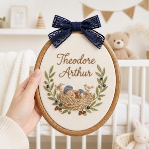 May include: An oval wooden embroidery hoop containing a cream fabric panel with the embroidered name Theodore Arthur. The design features a nest with a sleeping baby and a teddy bear, surrounded by green leaves, berries, and birds, topped with a navy blue lace bow.