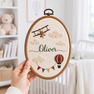 Può includere: Un telaio da ricamo ovale in legno contenente un pannello in tessuto color crema. Il design presenta un biplano marrone, una mongolfiera rossa e bianca, soffici nuvole bianche e bandierine colorate. Il nome Oliver è ricamato in corsivo verde scuro.