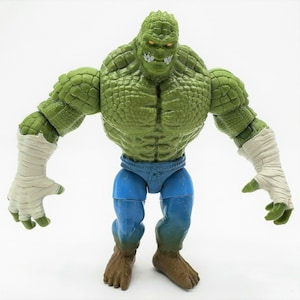 May include: A green action figure of a crocodile-like humanoid with a textured, scaly appearance. The figure has blue shorts, brown feet, and white bandages on its forearms. The figure has a menacing expression with yellow eyes.
