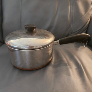 May include: A stainless steel saucepan with a lid and a black handle. The pot has a copper-coloured base and a black knob on the lid. The handle has a metal loop for hanging. The pot is on a grey surface.