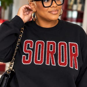 May include: A black long-sleeve sweatshirt featuring the word SOROR in large, bold letters. The letters are filled with red rhinestones and outlined with small white pearls. The sweatshirt has a crew neckline and a relaxed fit.