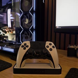 Könnte beinhalten: Ein weiß-schwarzer kabelloser Gamecontroller, der in einer passenden rechteckigen Ladestation ruht. Die Station zeigt die PlayStation-Symbole Dreieck, Kreis, Kreuz und Quadrat, die auf der Vorderseite in Schwarz und Weiß eingeprägt sind.