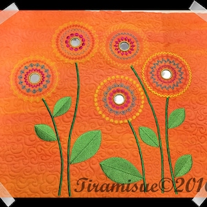 Puede incluir: Un tapiz de pared acolchado con fondo naranja y cinco flores bordadas. Las flores son amarillas, naranjas y verdes con un espejo en el centro de cada flor.