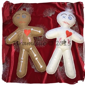 May include: Two voodoo dolls, one brown and one white, with red hearts and stitched-on details. The dolls are lying on a red velvet background with silver glitter.