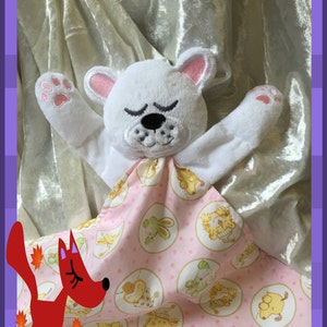 Pode incluir: Um urso de pelúcia branco com orelhas e patas cor-de-rosa está enrolado em um cobertor de bebê rosa e branco com um padrão de animais em círculos. O cobertor tem um fundo branco com bolinhas cor-de-rosa.