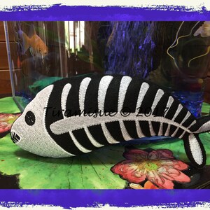 May include: A black and white plush fish with a skeleton design. The fish is lying on a green leaf with a pink flower in the background.