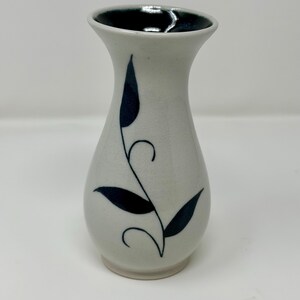 May include: A ceramic vase with a flared rim and a tapered neck. The exterior is off-white with a hand-painted dark blue leaf and vine motif. The interior of the vase is glazed in a deep, glossy dark blue, contrasting with the matte off-white body.