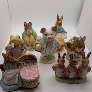 May include: A collection of seven ceramic animal figurines featuring characters in various poses. The set includes a pig in striped pajamas, a frog in a pink jacket, several rabbits in blue or red clothing, and a mouse with a basket of food.