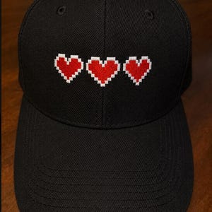 May include: A black baseball cap featuring an embroidered design of three red pixelated hearts arranged in a horizontal row across the front panel. The cap has a structured crown, a curved brim, and visible stitching details on the fabric surface.