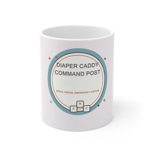 Op de afbeelding: Een witte keramische mok met een cirkelvormige afbeelding met een blauwe rand. De tekst luidt DIAPER CADDY COMMAND POST en WIPES, CREAM, EMERGENCY COFFEE. Onder de tekst staan drie kleine vierkante iconen met A, B en C, omringd door sterretjes.