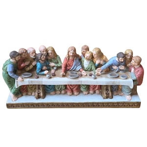 May include: A multi-colored figurine depicting the Last Supper, featuring thirteen figures seated at a long rectangular table covered with a white cloth. The figures wear robes in shades of red, blue, green, and brown, with small plates and cups.