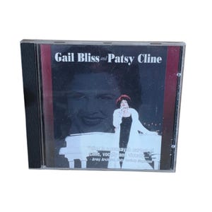 Può includere: Un CD musicale in una custodia di plastica trasparente con il titolo Gail Bliss and Patsy Cline. La copertina mostra un grande ritratto sbiadito dietro un'immagine più piccola di una persona con cappotto bianco e abito nero davanti a un piano.