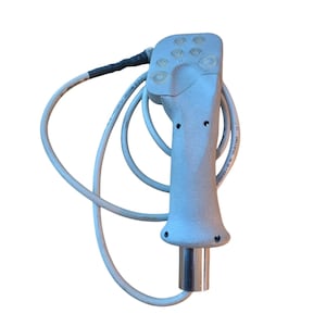 May include: A light gray handheld control device with a textured ergonomic grip, featuring six circular buttons on the top section. A long white cable is attached to the base of the handle, which also includes a metallic cylindrical connector at the bottom.