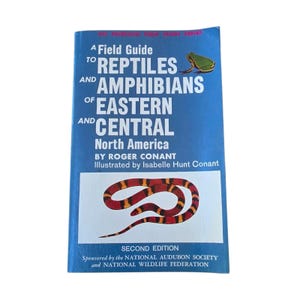 Może przedstawiać: Niebieska książka w miękkiej oprawie zatytułowana A Field Guide to Reptiles and Amphibians of Eastern and Central North America autorstwa Rogera Conanta. Okładka zawiera ilustrację zielonej żaby u góry oraz czerwono-czarno-żółtego węża u dołu.
