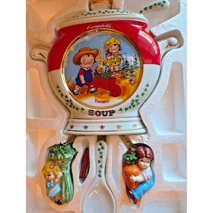 May include: A Campbell's Tomato Soup wall clock featuring a central illustration of two children in a garden with a red wheelbarrow. The clock has gold trim, ivy accents, and two hanging weights shaped like a child with celery and a child with a carrot.