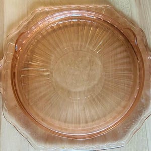 May include: A round, translucent pink glass serving plate featuring a central circular area with radiating linear ridges. The plate has a scalloped edge and two integrated side handles, showcasing a smooth, polished finish throughout the piece.