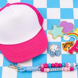 May include: A bright pink and white trucker hat with a mesh back, accompanied by a collection of colorful embroidered patches including a star, heart, rainbow, tiger, and letter P, plus a beaded keychain with the name PHOEBE and a heart icon.
