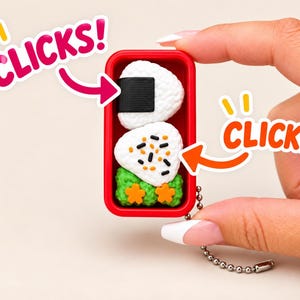 May include: A rectangular red plastic container featuring two white triangular rice balls, one wrapped in black seaweed and the other topped with black and orange sesame seeds, alongside green textured garnish with orange flower shapes and a metal chain.