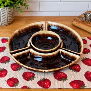 May include: A round ceramic serving dish with a dark brown glossy glaze and cream-colored edges. The dish features a central circular bowl surrounded by three separate wedge-shaped compartments, creating a multi-sectioned platter design.