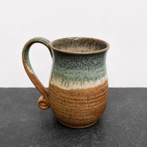 May include: A ceramic mug featuring a two-tone glaze with a speckled brown base and a mottled sage green upper section. The handle is curved with a decorative spiral detail at the bottom, and the rim shows a smooth, rounded edge finish.