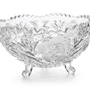 May include: A clear glass bowl featuring a scalloped rim and three small feet. The exterior surface is decorated with intricate pressed patterns, including prominent frosted rose motifs and starburst designs that catch the light.