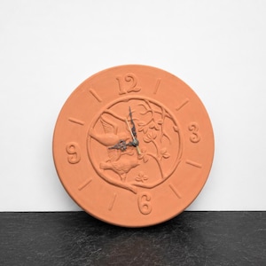 May include: A round terracotta wall clock featuring embossed numbers 3, 6, 9, and 12. The center displays a relief carving of two birds perched on a branch with leaves. The clock has thin, dark metal hour, minute, and second hands at the center.