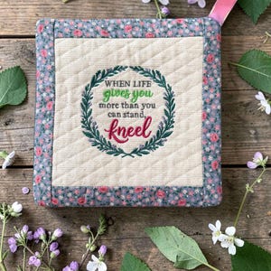 May include: A square quilted potholder with a cream-colored center featuring an embroidered green wreath and the text "WHEN LIFE gives you more than you can stand, kneel" in green and pink. It has a floral border and a pink fabric hanging loop.