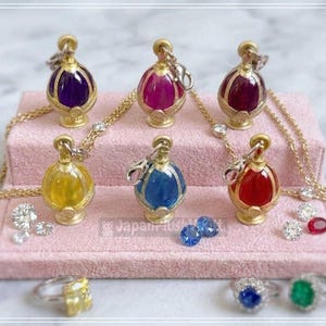 Könnte beinhalten: Sechs goldfarbene Anhänger in Form ovaler Flaschen mit farbigen Zentren in Lila, Pink, Dunkelrot, Gelb, Blau und Rot. Jeder Anhänger hat einen Karabinerverschluss und eine Goldkette. Lose facettierte Edelsteine und drei Ringe mit gelben, blauen und grünen Steinen.