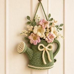 May include: A crocheted wall hanging shaped like a green watering can, featuring a cream-colored bow on the front and a bouquet of pink and white crocheted flowers with green leaves emerging from the top, suspended by a matching green yarn loop.