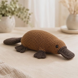 May include: A brown crocheted platypus toy featuring a dark brown bill, dark brown webbed feet, and a dark brown flat tail. The body is crafted from light brown yarn with a visible stitch texture and includes small black plastic eyes on the sides.