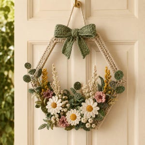 May include: A hexagonal crochet wreath featuring a cream-coloured frame topped with a sage green bow. The arrangement includes crocheted white daisies with yellow centres, small pink flowers, yellow sprigs, and various green eucalyptus-style leaves.