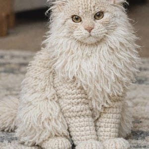 May include: A handcrafted cream-colored crocheted cat figurine featuring a dense, shaggy yarn texture around the neck and chest, a smooth crocheted body, pointed ears, and amber-colored eyes, sitting in an upright position with a long tail.