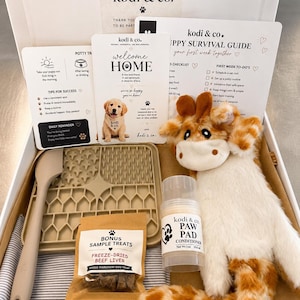 May include: A puppy care gift box from kodi & co. containing a beige silicone lick mat, a spatula, a bag of freeze-dried beef liver treats, a 1 oz paw pad conditioner stick, and a plush giraffe toy, alongside printed puppy survival guide cards.