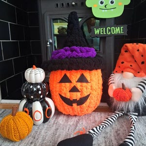 May include: A collection of Halloween decorations including a crocheted orange pumpkin wearing a black and purple witch hat, a black and white ceramic stack of pumpkins with the word BOO, a small orange knit pumpkin, and a gnome with a tall orange hat.