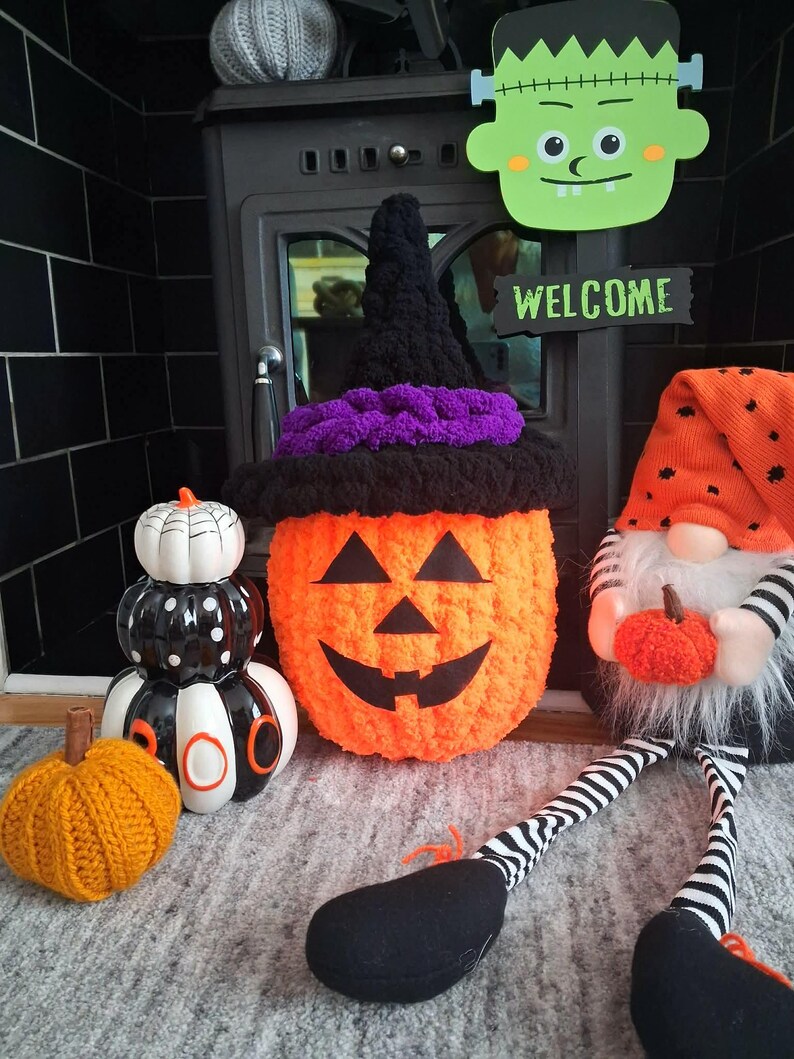 May include: A collection of Halloween decorations including a crocheted orange pumpkin wearing a black and purple witch hat, a black and white ceramic stack of pumpkins with the word BOO, a small orange knit pumpkin, and a gnome with a tall orange hat.