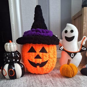 May include: A collection of Halloween decorations including a stacked ceramic pumpkin trio in black, white, and orange, a large orange crocheted pumpkin wearing a black and purple witch hat, a white plush ghost holding a BOO banner, and a small orange crocheted pumpkin.