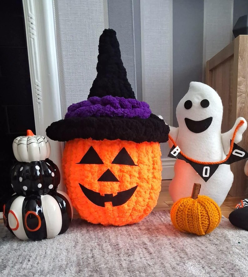 May include: A collection of Halloween decorations including a stacked ceramic pumpkin trio in black, white, and orange, a large orange crocheted pumpkin wearing a black and purple witch hat, a white plush ghost holding a BOO banner, and a small orange crocheted pumpkin.