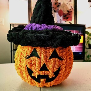 May include: A round, orange, crocheted pumpkin plush toy featuring a black felt jack-o'-lantern face with eyelashes. The pumpkin wears a tall, black, crocheted witch hat with a thick, textured purple band around the base of the pointed crown.