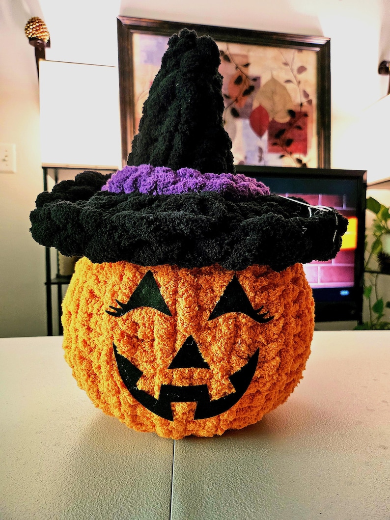 May include: A round, orange, crocheted pumpkin plush toy featuring a black felt jack-o'-lantern face with eyelashes. The pumpkin wears a tall, black, crocheted witch hat with a thick, textured purple band around the base of the pointed crown.